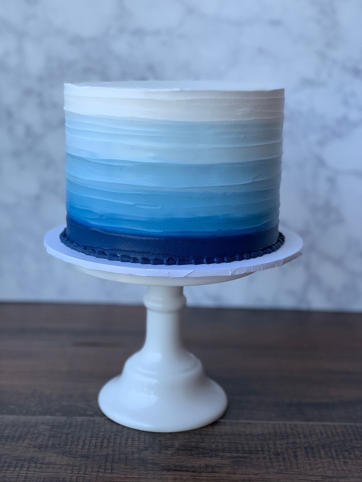 Blue gradient buttercream cake on a white pedestal stand against marble background