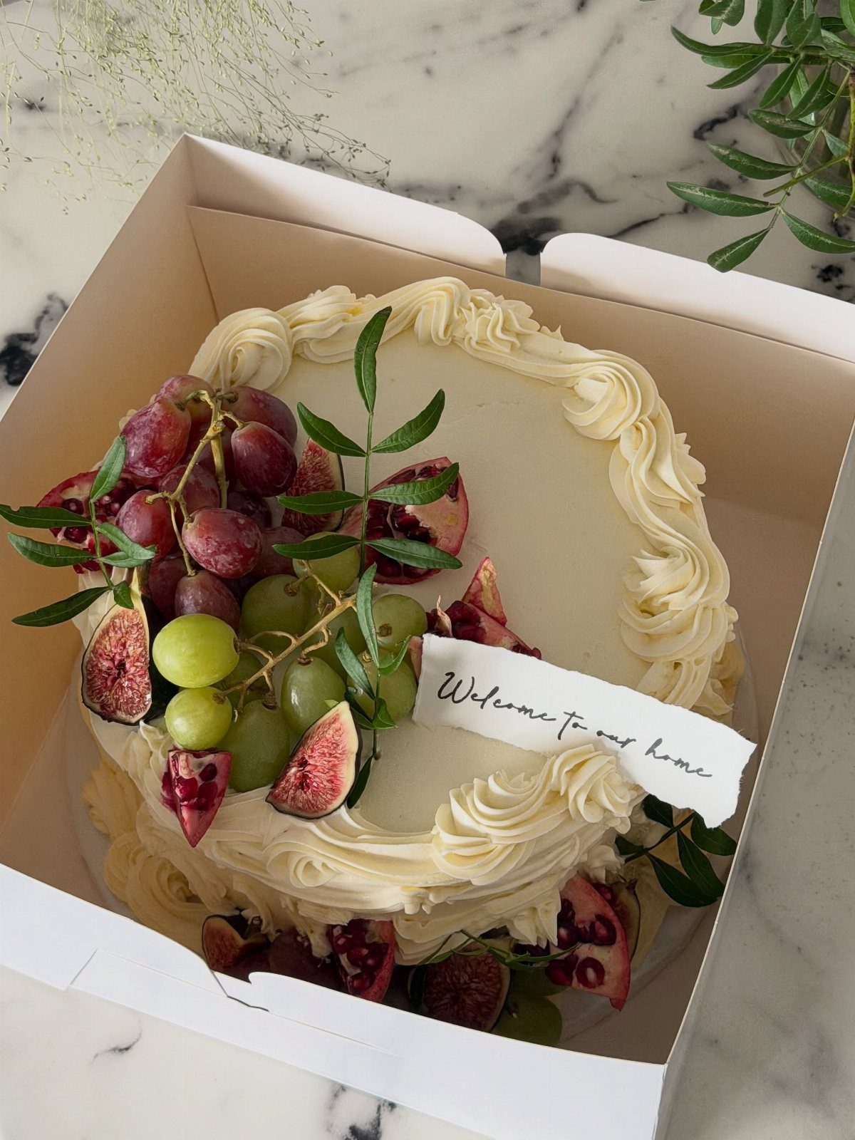 White frosted cake decorated with fresh grapes, sliced figs, and a welcome home note.