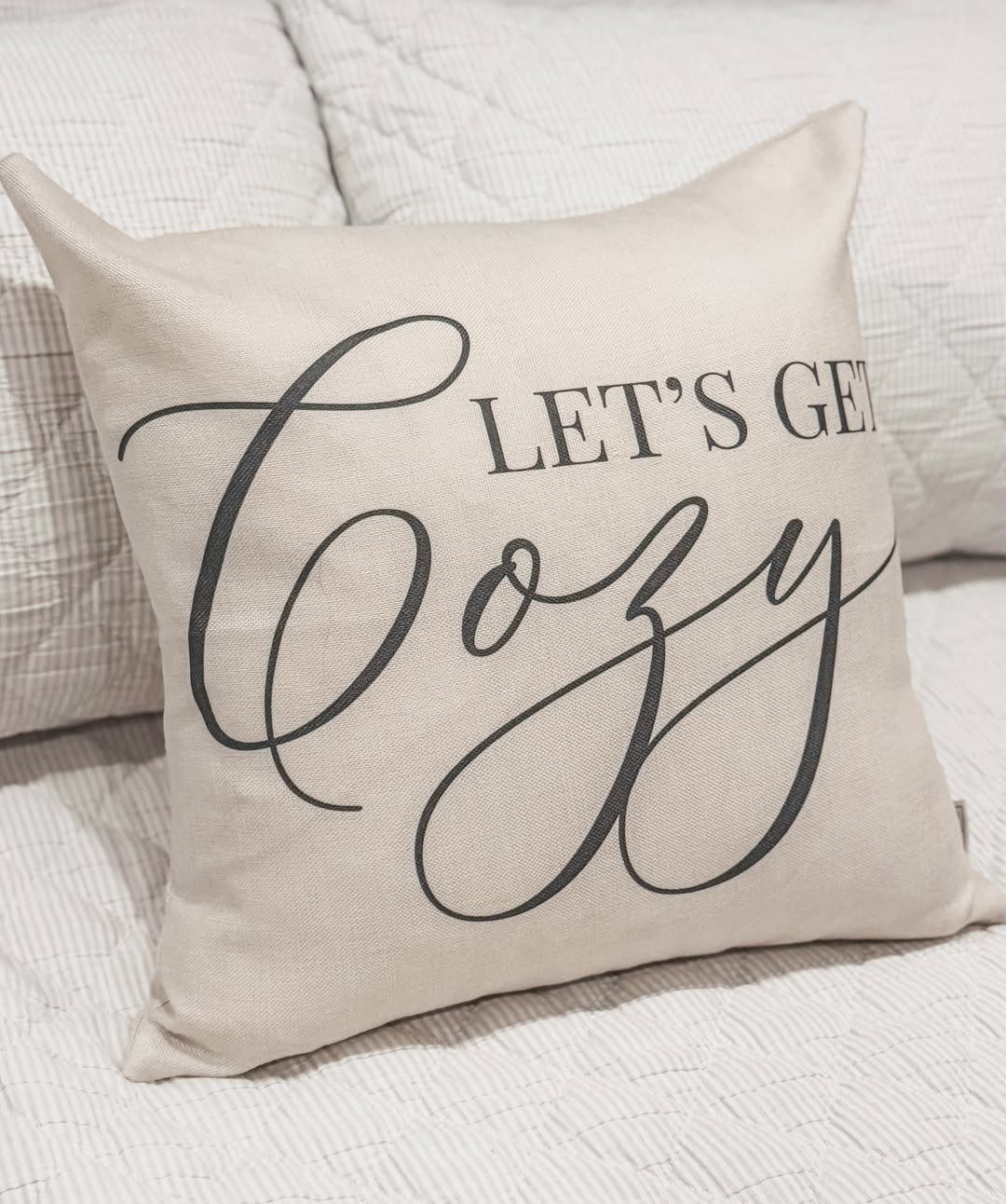 Beige decorative pillow with 'LET'S GET Cozy' text on a white quilted bed.