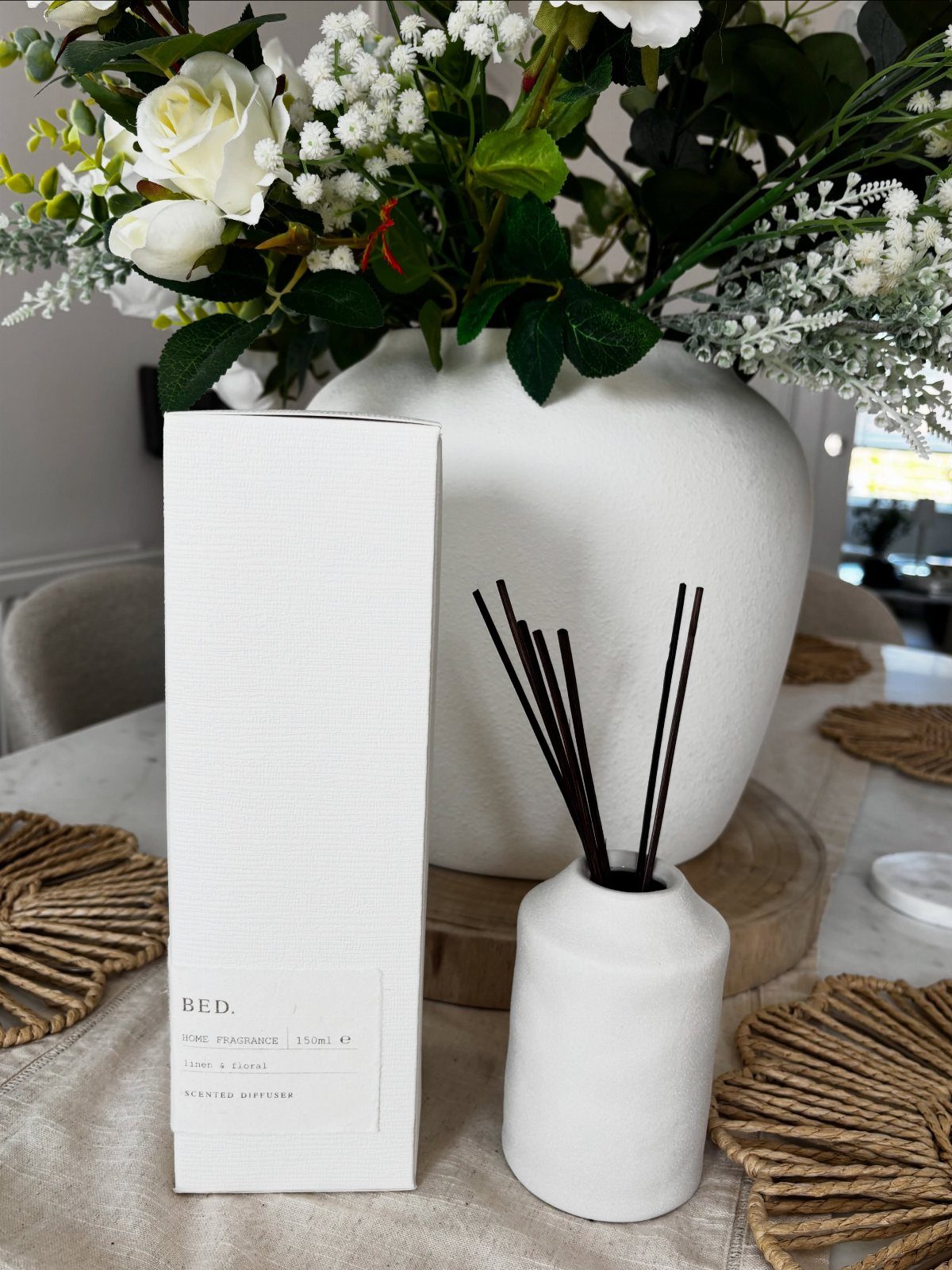 White ceramic reed diffuser and packaging next to a vase of white roses.