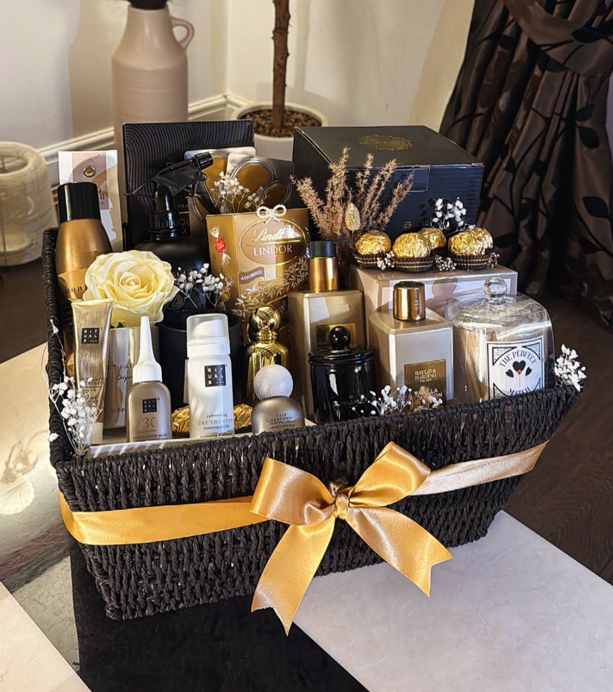 Black woven gift basket with gold ribbon containing perfumes, skincare, and chocolates.