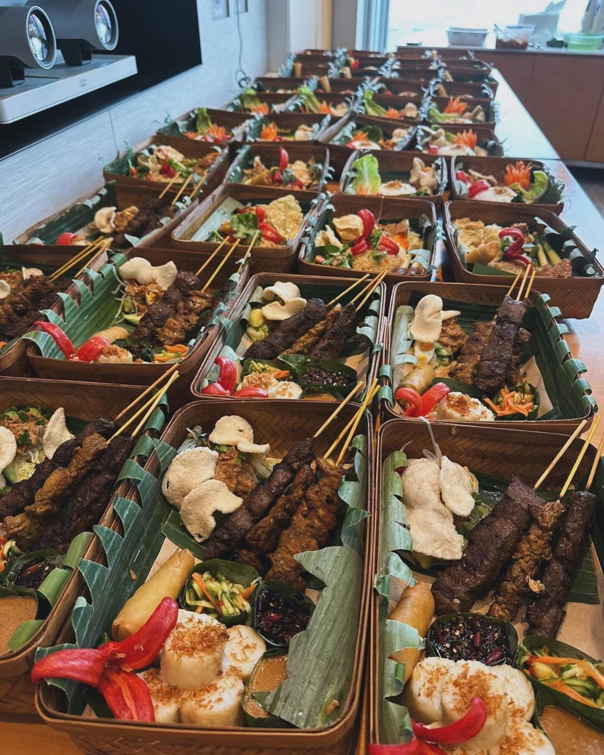 Grilled meat skewers and rice cakes in banana leaf lined meal boxes.