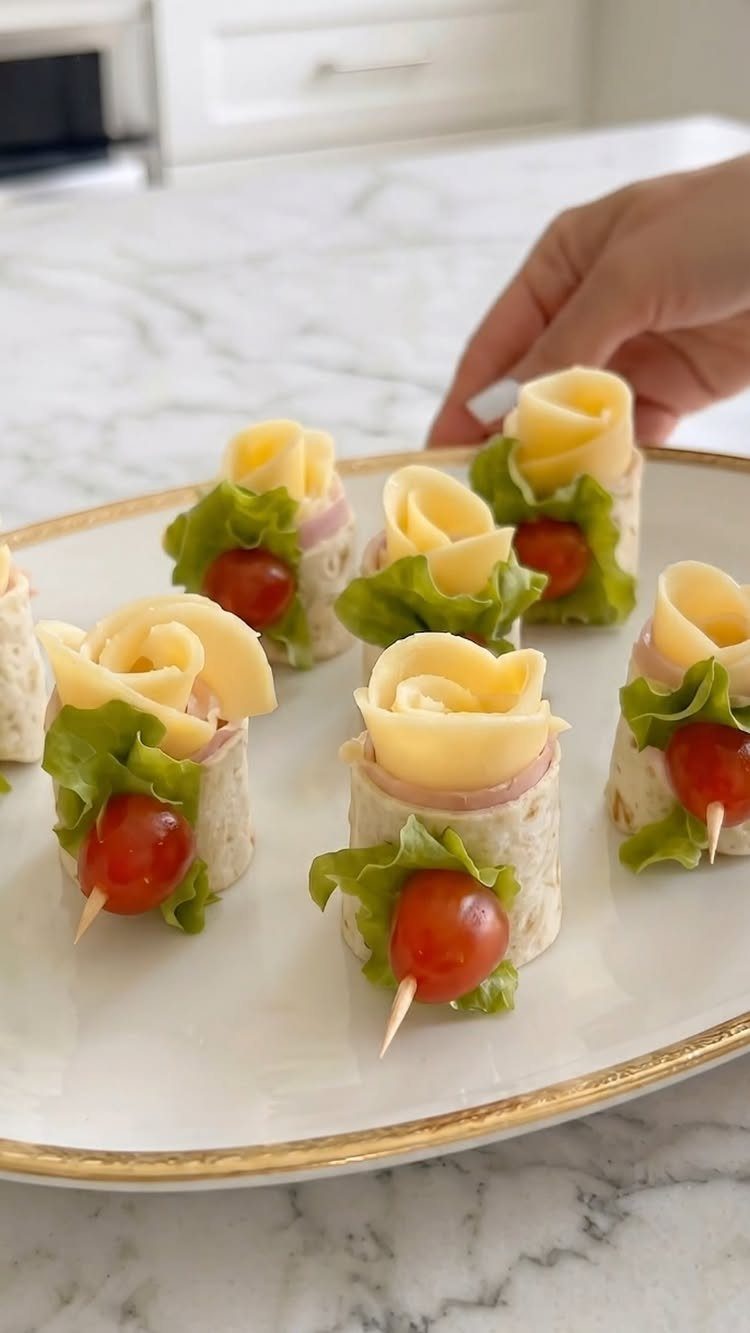 Bite-sized tortilla wraps with cheese roses and cherry tomatoes on a white plate.